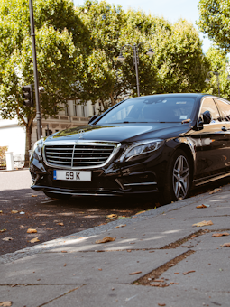 A sleek black luxury sedan is parked on a city street with green leafy trees in the background. The car is angled slightly away from the curb, and dry leaves are scattered on the ground around it. The vehicle features a distinctive front grille and has a European license plate.