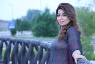 A woman with long, wavy brown hair is smiling slightly while leaning against a decorative railing. The background is a blurred outdoor scene with green trees and a street sign visible.