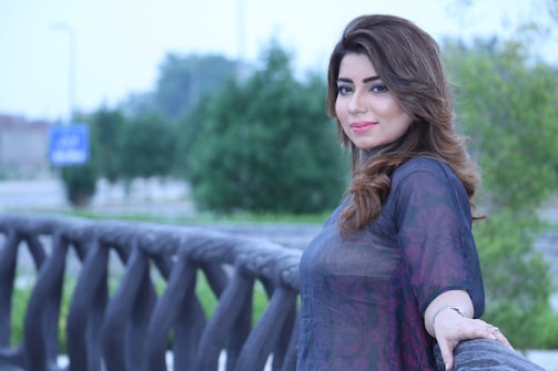 A woman with long, wavy brown hair is smiling slightly while leaning against a decorative railing. The background is a blurred outdoor scene with green trees and a street sign visible.