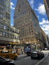 A city street scene featuring a tall, brick skyscraper under a partly cloudy blue sky. The foreground includes a yellow NYC taxi and a delivery truck near a Wells Fargo branch. Pedestrians are visible on the sidewalk alongside parked cars.