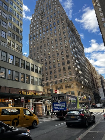 A city street scene featuring a tall, brick skyscraper under a partly cloudy blue sky. The foreground includes a yellow NYC taxi and a delivery truck near a Wells Fargo branch. Pedestrians are visible on the sidewalk alongside parked cars.