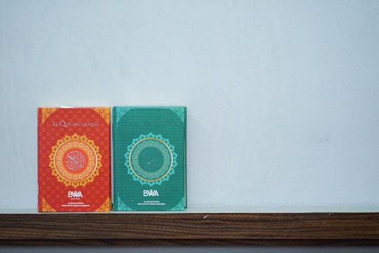 Two books with intricate designs rest on a wooden ledge against a light gray wall. The left book has a red cover with golden circular patterns and Arabic text, while the right book has a green cover with similar decorative elements.