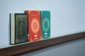 Three books are neatly placed on a wooden shelf. The first book has a dark cover with ornate gold detailing, suggesting a religious text. The middle book is red with intricate circular patterns and some visible text. The third book is green with similar design elements as the second book.