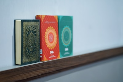 Three books are neatly placed on a wooden shelf. The first book has a dark cover with ornate gold detailing, suggesting a religious text. The middle book is red with intricate circular patterns and some visible text. The third book is green with similar design elements as the second book.