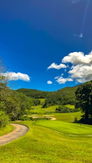 A lush green golf course with neatly trimmed grass sits under a bright blue sky. Fluffy white clouds float above, and the course is surrounded by wooded hills.