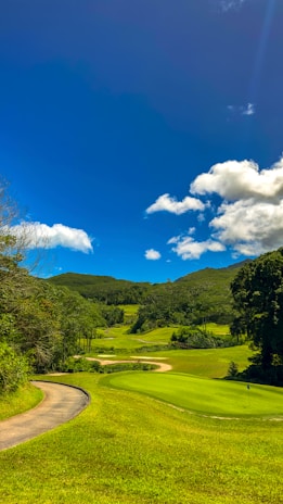 A lush green golf course with neatly trimmed grass sits under a bright blue sky. Fluffy white clouds float above, and the course is surrounded by wooded hills.