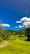 A lush green golf course with neatly trimmed grass sits under a bright blue sky. Fluffy white clouds float above, and the course is surrounded by wooded hills.
