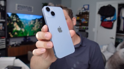 A person holds a light blue smartphone with the Apple logo prominently displayed. The background features a television displaying a vibrant nature scene with green trees and cliffs.
