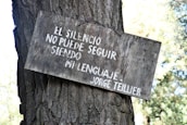 A wooden sign with white Spanish text is attached to the trunk of a tree. The text reads 'EL SILENCIO NO PUEDE SEGUIR SIENDO MI LENGUAJE - JORGE TEILLIER.' The sign appears weathered and the tree trunk is textured with rough bark. The background features out-of-focus foliage.