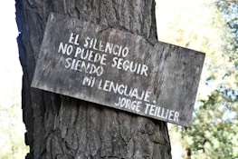 A wooden sign with white Spanish text is attached to the trunk of a tree. The text reads 'EL SILENCIO NO PUEDE SEGUIR SIENDO MI LENGUAJE - JORGE TEILLIER.' The sign appears weathered and the tree trunk is textured with rough bark. The background features out-of-focus foliage.