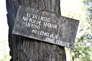 A wooden sign with white Spanish text is attached to the trunk of a tree. The text reads 'EL SILENCIO NO PUEDE SEGUIR SIENDO MI LENGUAJE - JORGE TEILLIER.' The sign appears weathered and the tree trunk is textured with rough bark. The background features out-of-focus foliage.
