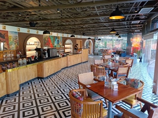 A well-lit cafe interior with a long wooden counter on the left side lined with various coffee machines and jars. The ceiling has a decorative design with black hanging pendant lights evenly spaced. The floor features a bold black and white geometric pattern. On the right side, there are several wooden tables and chairs, each with colorful cushions. Large glass windows at the far end allow natural light to enter, and some potted plants are visible near the windows.