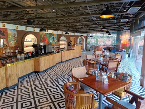 A well-lit cafe interior with a long wooden counter on the left side lined with various coffee machines and jars. The ceiling has a decorative design with black hanging pendant lights evenly spaced. The floor features a bold black and white geometric pattern. On the right side, there are several wooden tables and chairs, each with colorful cushions. Large glass windows at the far end allow natural light to enter, and some potted plants are visible near the windows.