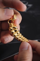 A close-up of hands holding a shiny gold pendant shaped like a spiral seashell. The intricate design features reflective, smooth surfaces and a detailed top loop for attaching to a chain.