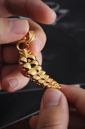 A close-up of hands holding a shiny gold pendant shaped like a spiral seashell. The intricate design features reflective, smooth surfaces and a detailed top loop for attaching to a chain.