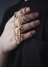 A hand gently holds a gold bullet-shaped pendant attached to a beaded necklace. The hand has neatly manicured nails and rests against a dark fabric background, highlighting the shiny and elegant design of the jewelry.