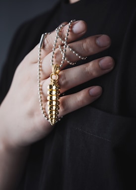 A hand gently holds a gold bullet-shaped pendant attached to a beaded necklace. The hand has neatly manicured nails and rests against a dark fabric background, highlighting the shiny and elegant design of the jewelry.