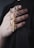 A hand gently holds a gold bullet-shaped pendant attached to a beaded necklace. The hand has neatly manicured nails and rests against a dark fabric background, highlighting the shiny and elegant design of the jewelry.
