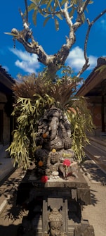 An intricately carved stone sculpture of Ganesha is surrounded by vibrant greenery and flowers. The statue is set against a bright blue sky with a few fluffy clouds. A tree with broad leaves extends upwards behind the statue, creating a natural frame. A small offering of various colored flowers is placed on a platform in front of the sculpture, likely part of a religious or cultural ritual, and a second smaller statue rests at the base, possibly depicting Buddha.