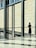 A person in a black outfit stands alone in a modern, spacious, and well-lit architectural space. Large glass windows and a wall with a geometric pattern dominate the scene. The person appears to be deep in thought or contemplation.