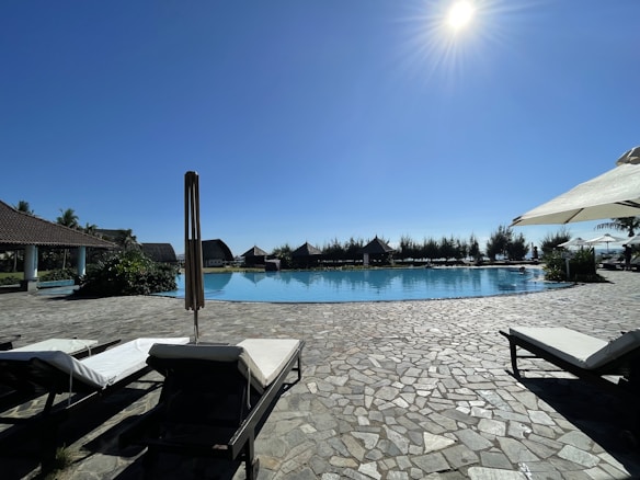 A sunlit swimming pool is surrounded by lounge chairs with white cushions and large umbrellas providing shade. The pool area is paved with stone tiles, and there are several thatched-roof structures in the background. Tall trees line the horizon under a clear blue sky with the sun shining brightly.