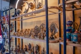 A collection of intricately crafted sculptures and figurines is displayed on glass shelves. The items include various traditional and cultural artifacts, such as mini statues of deities and decorative pots. The pieces are predominantly metallic with some colorful elements visible, and the setup appears to have a cultural and historical significance.