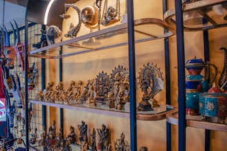 A collection of intricately crafted sculptures and figurines is displayed on glass shelves. The items include various traditional and cultural artifacts, such as mini statues of deities and decorative pots. The pieces are predominantly metallic with some colorful elements visible, and the setup appears to have a cultural and historical significance.