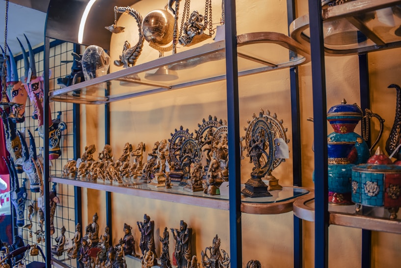 A collection of intricately crafted sculptures and figurines is displayed on glass shelves. The items include various traditional and cultural artifacts, such as mini statues of deities and decorative pots. The pieces are predominantly metallic with some colorful elements visible, and the setup appears to have a cultural and historical significance.