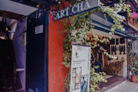 An art gallery entrance adorned with plants and a variety of sculptures and artifacts displayed inside. A large banner advertises a current art exhibition, and signage for the gallery is visible. The setting includes a vibrant mix of colors and textures.