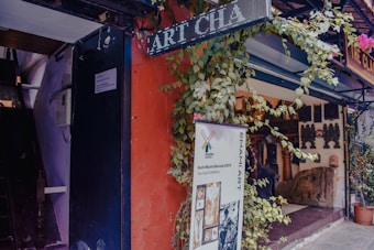 An art gallery entrance adorned with plants and a variety of sculptures and artifacts displayed inside. A large banner advertises a current art exhibition, and signage for the gallery is visible. The setting includes a vibrant mix of colors and textures.