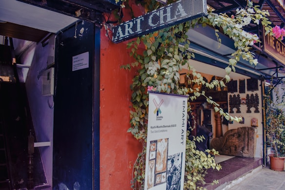 An art gallery entrance adorned with plants and a variety of sculptures and artifacts displayed inside. A large banner advertises a current art exhibition, and signage for the gallery is visible. The setting includes a vibrant mix of colors and textures.