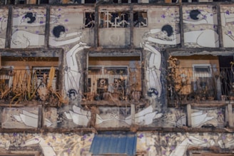 A building facade featuring artwork depicting numerous abstract human figures interacting in various poses. The building appears to be old and worn, with some sections showing plants and decay. The artwork is monochromatic with occasional purple accents.