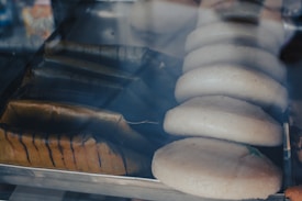 A display case contains rows of round, light-colored discs resembling bread or pastries. Next to them are rectangular food items wrapped in leaves, likely tamales.