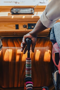 A hand firmly grips the gear shift of a vehicle. The dashboard features a radio and a few control switches mounted on a wooden panel. The gear shift is decorated with multiple colored bands, adding a personal touch to the vehicle's interior.