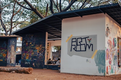 Outdoor art space featuring walls covered with colorful graffiti and murals. Two structures with flat roofs and open sides provide a casual setting. In the foreground, a large log lies on the ground. People are seated under one of the structures engaged in activities.