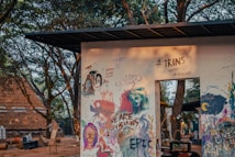 A wall adorned with colorful graffiti and murals, featuring various artistic expressions including faces, abstract patterns, and writings. The surrounding environment includes trees and rustic outdoor elements.