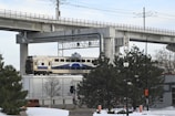 A commuter train travels on a track elevated above the snowy landscape. The foreground features snow-covered ground with trees and a street lamp. Overhead, concrete structures of another elevated track or bridge are visible. An orange vehicle and traffic cones are partially obscured by the trees.