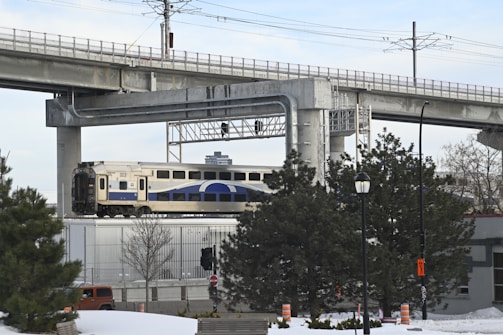 A commuter train travels on a track elevated above the snowy landscape. The foreground features snow-covered ground with trees and a street lamp. Overhead, concrete structures of another elevated track or bridge are visible. An orange vehicle and traffic cones are partially obscured by the trees.