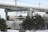 A commuter train travels on a track elevated above the snowy landscape. The foreground features snow-covered ground with trees and a street lamp. Overhead, concrete structures of another elevated track or bridge are visible. An orange vehicle and traffic cones are partially obscured by the trees.