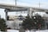 A commuter train travels on a track elevated above the snowy landscape. The foreground features snow-covered ground with trees and a street lamp. Overhead, concrete structures of another elevated track or bridge are visible. An orange vehicle and traffic cones are partially obscured by the trees.