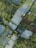 Aerial view of a patchwork of agricultural fields with varying shades of green and brown, interspersed with trees and shrubs. Long rectangular plots are covered with plastic sheets, possibly for greenhouse or protective farming. There are a few small structures, possibly farmhouses or sheds, scattered throughout the landscape. Narrow pathways and roads weave between the fields, providing access.