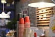 A stack of disposable cups in red, black, and cream colors is organized neatly on a countertop. The background features a cozy cafe interior with a stone wall and wooden shelves. Warm lighting hangs overhead, contributing to the inviting atmosphere.