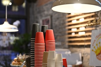 A stack of disposable cups in red, black, and cream colors is organized neatly on a countertop. The background features a cozy cafe interior with a stone wall and wooden shelves. Warm lighting hangs overhead, contributing to the inviting atmosphere.