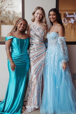 Three women are standing together, each wearing an elegant gown. The woman on the left wears a turquoise satin dress with off-the-shoulder sleeves. The woman in the middle is dressed in a sparkling gown with sequins and stripes in shades of silver and turquoise. The woman on the right is wearing a light blue gown with lace and sheer sleeves. They are all smiling and appear to be at a formal event.