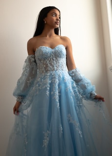 A woman wearing an elegant, strapless blue gown with intricate lace and embroidery details. The dress features sheer sleeves and a flowing skirt. She is poised with an upward gaze and stands against a softly lit neutral-colored background.
