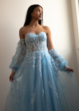 A woman wearing an elegant, strapless blue gown with intricate lace and embroidery details. The dress features sheer sleeves and a flowing skirt. She is poised with an upward gaze and stands against a softly lit neutral-colored background.