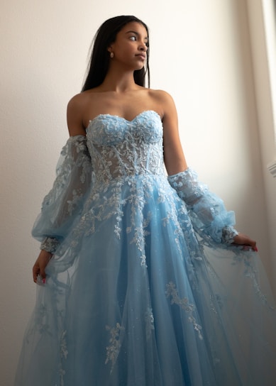 A woman wearing an elegant, strapless blue gown with intricate lace and embroidery details. The dress features sheer sleeves and a flowing skirt. She is poised with an upward gaze and stands against a softly lit neutral-colored background.