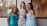 Three women in elegant evening gowns are smiling and posing together in front of a window. One wears a turquoise off-shoulder dress, the second wears a sequined gown with diagonal stripes, and the third wears a light blue dress with floral lace details. The background shows an indoor setting with a hint of wedding dresses and a sign.