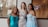 Three women in elegant evening gowns are smiling and posing together in front of a window. One wears a turquoise off-shoulder dress, the second wears a sequined gown with diagonal stripes, and the third wears a light blue dress with floral lace details. The background shows an indoor setting with a hint of wedding dresses and a sign.