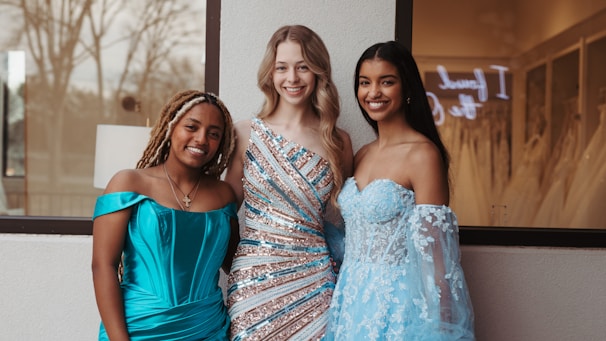Three women in elegant evening gowns are smiling and posing together in front of a window. One wears a turquoise off-shoulder dress, the second wears a sequined gown with diagonal stripes, and the third wears a light blue dress with floral lace details. The background shows an indoor setting with a hint of wedding dresses and a sign.